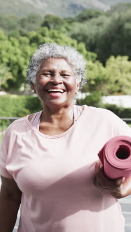 Vertical-video-portrait-of-happy-senior-african-american-woman-with-yoga-mat-in-garden,-slow-motion