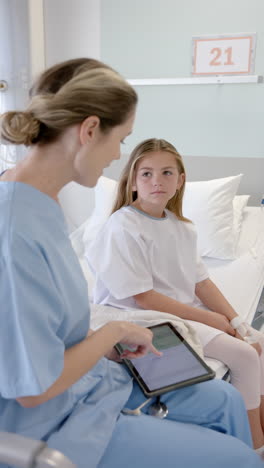 Vertical-video-of-caucasian-female-doctor-with-tablet-talking-with-girl-patient-on-bed,-slow-motion