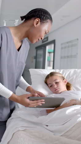 Vertical-video-of-diverse-doctor-and-girl-patient-lying-on-bed-using-tablet-in-hospital,-slow-motion