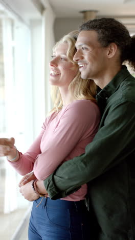 Video-Vertical-De-Una-Feliz-Pareja-Diversa-Abrazándose-En-La-Ventana-De-Casa,-Cámara-Lenta