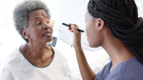 African-american-female-doctor-testing-eyes-of-senior-female-patient-in-hospital-room,-slow-motion