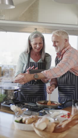 Vertical-video-of-happy-senior-caucasian-couple-cooking-dinner,-using-tablet-in-kitchen,-slow-motion