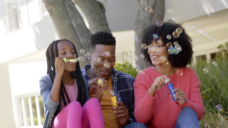 Felices-Padres-Afroamericanos-Con-Su-Hija-Soplando-Burbujas-En-El-Jardín-De-Casa,-Cámara-Lenta