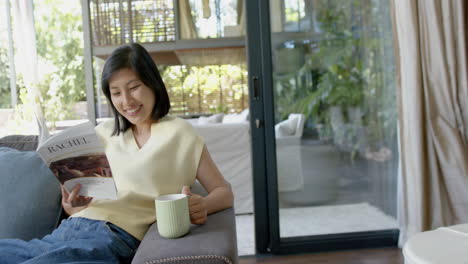 Feliz-Mujer-Asiática-Sentada-En-El-Sofá-Con-Una-Taza-Y-Leyendo-Un-Libro-En-Una-Soleada-Sala-De-Estar,-Cámara-Lenta