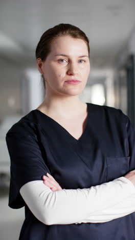 Vertical-video-of-portrait-of-happy-caucasian-female-doctor-wearing-scrubs-in-hospital,-slow-motion