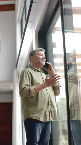 Video-Vertical-De-Un-Feliz-Hombre-Birracial-Mayor-Hablando-En-Un-Teléfono-Inteligente-Junto-A-La-Ventana-En-Casa,-Cámara-Lenta