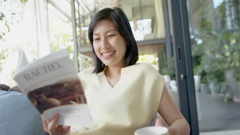 Happy-asian-woman-sitting-on-couch-with-cup-and-reading-book-in-sunny-living-room,-slow-motion