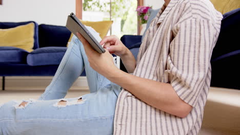 Midsection-of-biracial-man-sitting-on-floor-using-tablet-in-living-room,-slow-motion