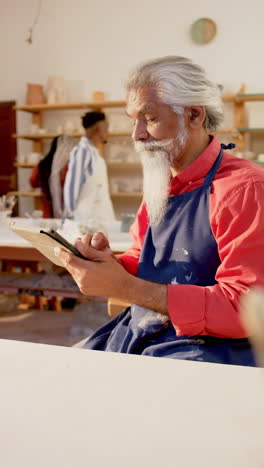 Focused-biracial-potter-with-long-beard-using-tablet-in-pottery-studio,-slow-motion