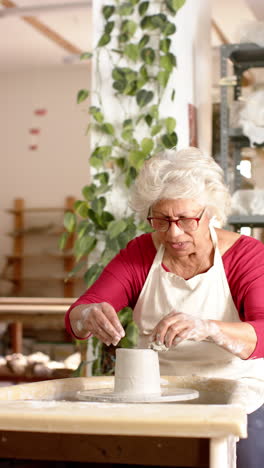 Focused-biracial-female-potter-with-gray-hair-working-on-clay-vase-in-pottery-studio,-slow-motion