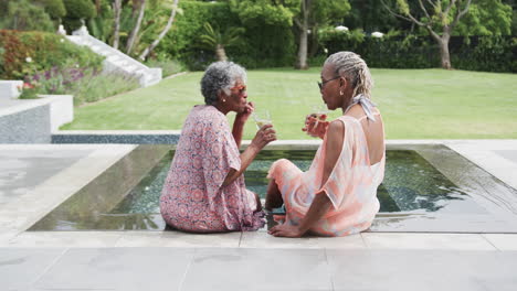 Happy-senior-african-american-female-friends-with-drinks-talking-by-pool,-copy-space,-slow-motion