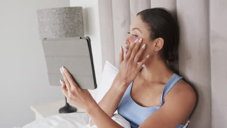 Biracial-woman-with-under-eye-patches-sitting-on-bed-using-tablet,-slow-motion