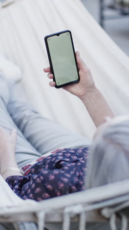 Video-Vertical-De-Una-Mujer-Caucásica-Mayor-Usando-Un-Teléfono-Inteligente-Con-Espacio-Para-Copiar-En-Hamaca,-Cámara-Lenta