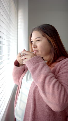 Vertical-video-of-plus-size-biracial-woman-drinking-tea-looking-out-of-window,-slow-motion