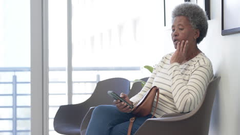 Mujer-Mayor-Afroamericana-Usando-Un-Teléfono-Inteligente-En-La-Sala-De-Espera-Del-Hospital,-Cámara-Lenta