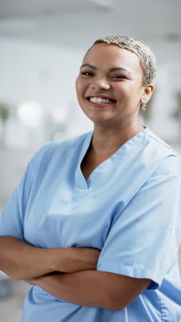 Vertical-video-of-portrait-of-happy-biracial-female-doctor-wearing-scrubs-in-hospital,-slow-motion