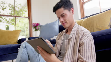 Happy-biracial-man-sitting-on-floor-using-tablet-in-living-room,-slow-motion