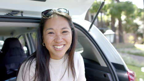 Feliz-Mujer-Asiática-Con-Gafas-De-Sol-Sentada-En-El-Baúl-Del-Auto-Y-Sonriendo-En-Un-Día-Soleado,-Cámara-Lenta