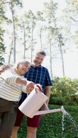 Happy-biracial-senior-couple-watering-plants-in-sunny-garden,-vertical