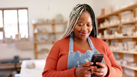 Glückliche-Afroamerikanische-Töpferin-Mit-Smartphone-Und-Lächeln-Im-Töpferstudio,-Zeitlupe