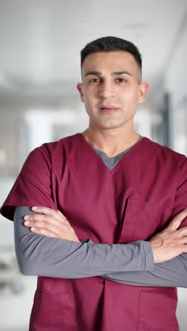 Vertical-video-of-portrait-of-happy-biracial-male-doctor-wearing-scrubs-in-hospital,-slow-motion