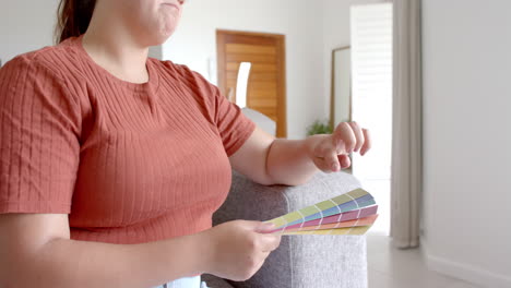 Thoughtful-plus-size-biracial-woman-on-couch-looking-at-paint-charts-in-living-room,-slow-motion