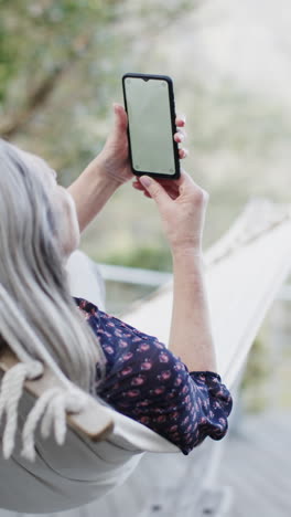 Video-Vertical-De-Una-Mujer-Caucásica-Mayor-Usando-Un-Teléfono-Inteligente-Con-Espacio-Para-Copiar-En-Hamaca,-Cámara-Lenta