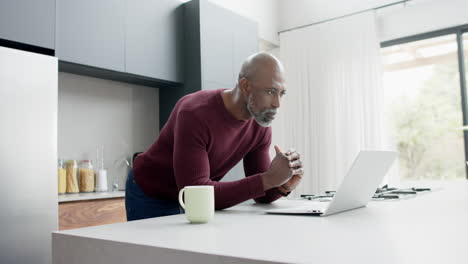 Glücklicher-Reifer-Mann-Mit-Gemischter-Abstammung-Mit-Einer-Tasse-Kaffee-Am-Laptop-In-Der-Küche,-Zeitlupe