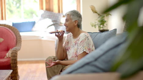 Feliz-Mujer-Birracial-Mayor-Sentada-En-El-Sofá-Y-Hablando-Por-Teléfono-Inteligente-En-Casa,-Cámara-Lenta