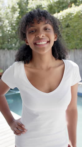 Video-Vertical-De-Una-Mujer-Birracial-Feliz-Con-Camiseta-Blanca,-Sonriendo-En-Un-Jardín-Soleado,-Cámara-Lenta