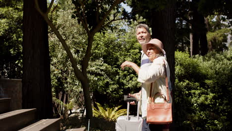 Happy-diverse-senior-couple-walking-with-luggage-to-a-house-in-sunny-outdoors