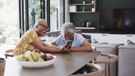 Amigas-Afroamericanas-Mayores-Usando-Smartphone-Y-Riéndose-En-El-Comedor,-Cámara-Lenta