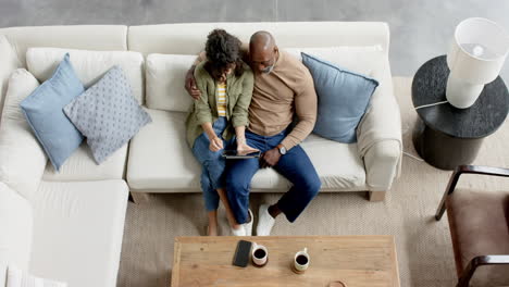 Happy-biracial-couple-sitting-on-couch-and-using-tablet-at-home,-slow-motion