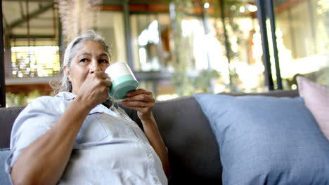 Mujer-Mayor-Birracial-Sentada-En-El-Sofá,-Bebiendo-Té-Y-Mirando-Hacia-El-Lado-De-Casa