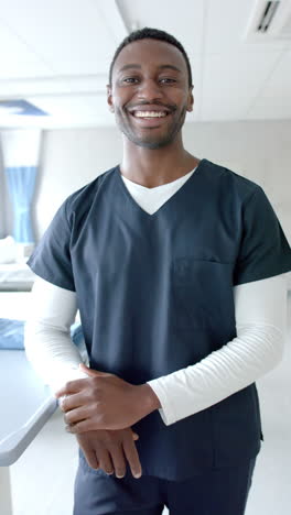 Vertical-video-portrait-of-smiling-african-american-male-doctor-in-scrubs-at-hospital,-slow-motion
