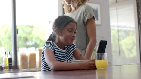 Feliz-Hija-Birracial-Usando-Un-Teléfono-Inteligente-En-Una-Cocina-Soleada-Con-Su-Madre,-Cámara-Lenta