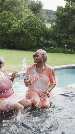 Video-Vertical-De-Amigas-Afroamericanas-Mayores-Sentadas-En-La-Piscina-Con-Bebidas,-Cámara-Lenta