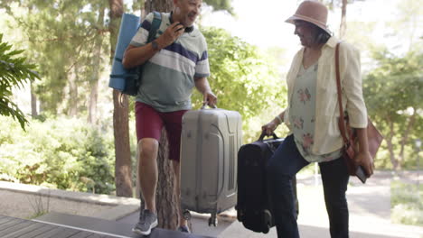 Happy-diverse-senior-couple-walking-with-luggage-to-a-house-in-sunny-outdoors