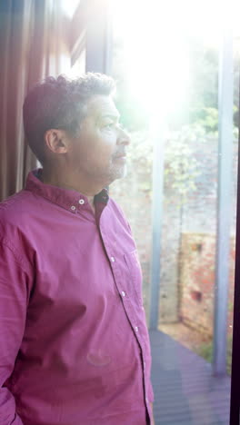 Portrait-of-focused-biracial-senior-man-leaning-and-looking-through-window-at-home,-vertical