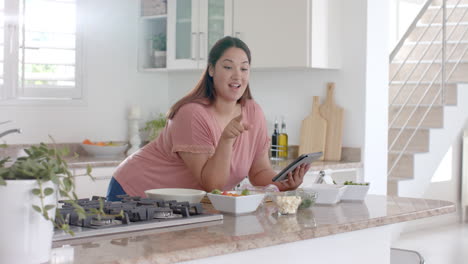 Happy-plus-size-biracial-woman-making-meal-with-vegetables-in-kitchen-and-using-tablet,-slow-motion