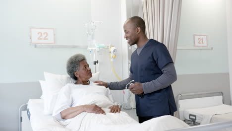 Happy-african-american-male-doctor-talking-with-female-senior-patient-in-hospital-room,-slow-motion