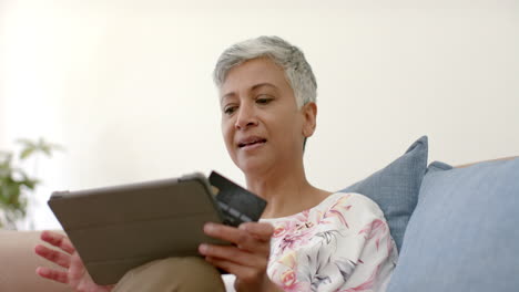 Feliz-Mujer-Birracial-Mayor-En-El-Sofá-Sosteniendo-Tarjeta-De-Crédito-Y-Usando-Tableta-En-Casa,-Cámara-Lenta