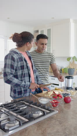 Vertical-video-of-happy-diverse-gay-male-couple-making-fruit-smoothie-in-kitchen,-slow-motion