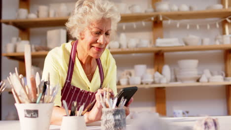 Happy-biracial-female-potter-with-gray-hair-using-smartphone-in-pottery-studio,-slow-motion