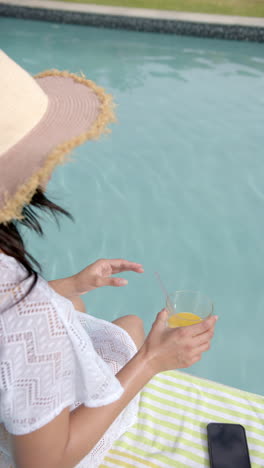 Vertical-video-of-biracial-teenage-girl-in-sunhat-sitting-by-sunny-pool-with-juice,-slow-motion