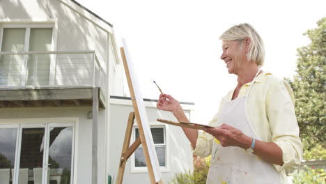 Feliz-Mujer-Mayor-Caucásica-Pintando-Cuadros-Y-Sonriendo-En-Un-Jardín-Soleado,-Cámara-Lenta,-Espacio-De-Copia