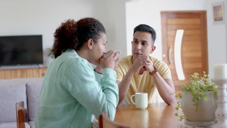 Happy-diverse-gay-male-couple-at-dining-table,-having-coffee-and-talking-at-home,-slow-motion