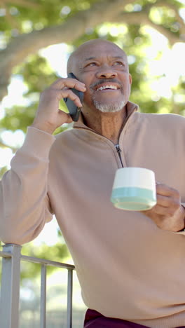 Vertical-video-of-happy-senior-biracial-man-talking-on-smartphone-at-balcony-at-home,-slow-motion