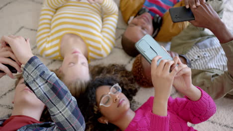 Happy-diverse-group-of-teenage-friends-using-smartphones-and-lying-on-carpet-at-home,-slow-motion