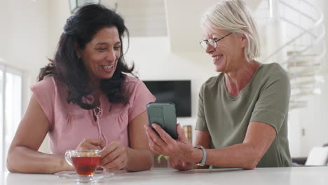 Felices-Y-Diversas-Mujeres-Mayores-Usando-Teléfonos-Inteligentes-Y-Discutiendo-En-Una-Cocina-Soleada,-Cámara-Lenta